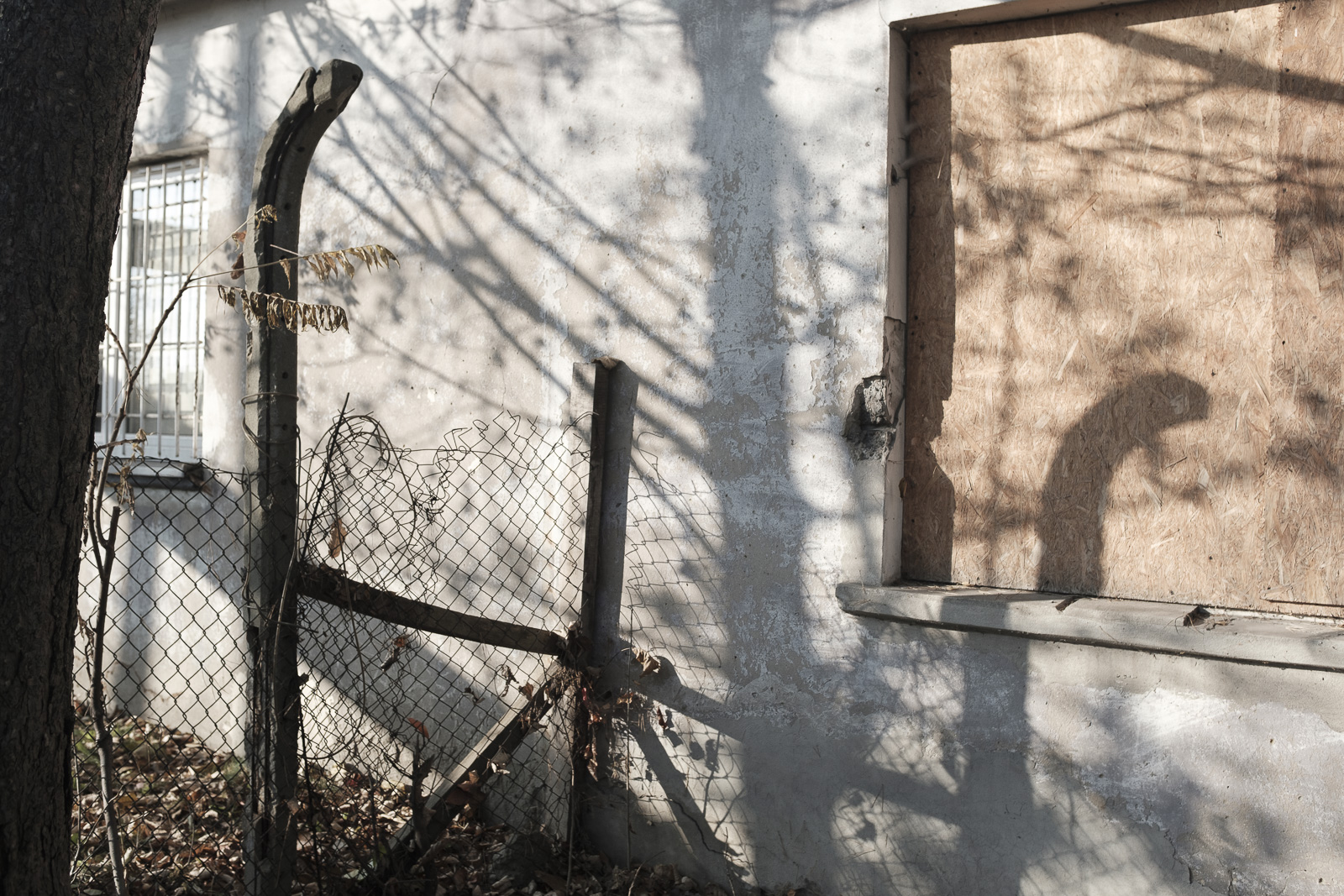 A boarded-up doorway and broken wire fence at the former Łagisza subcamp, with fence post shadows cast across the concrete wall.
