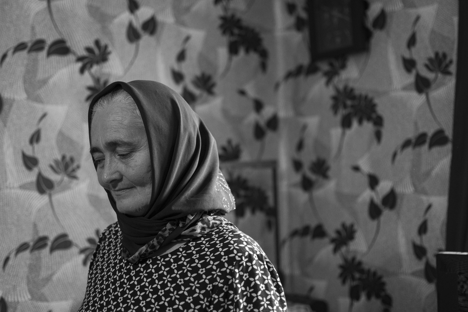Woman wearing a headscarf and patterned dress standing indoors against a floral wallpaper background with eyes closed