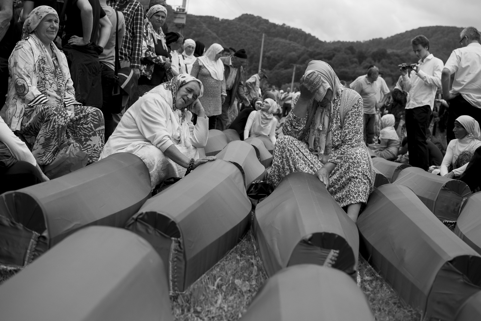 Women in headscarves sitting and mourning beside rows of coffin-shaped objects at an outdoor memorial site with hills in the background